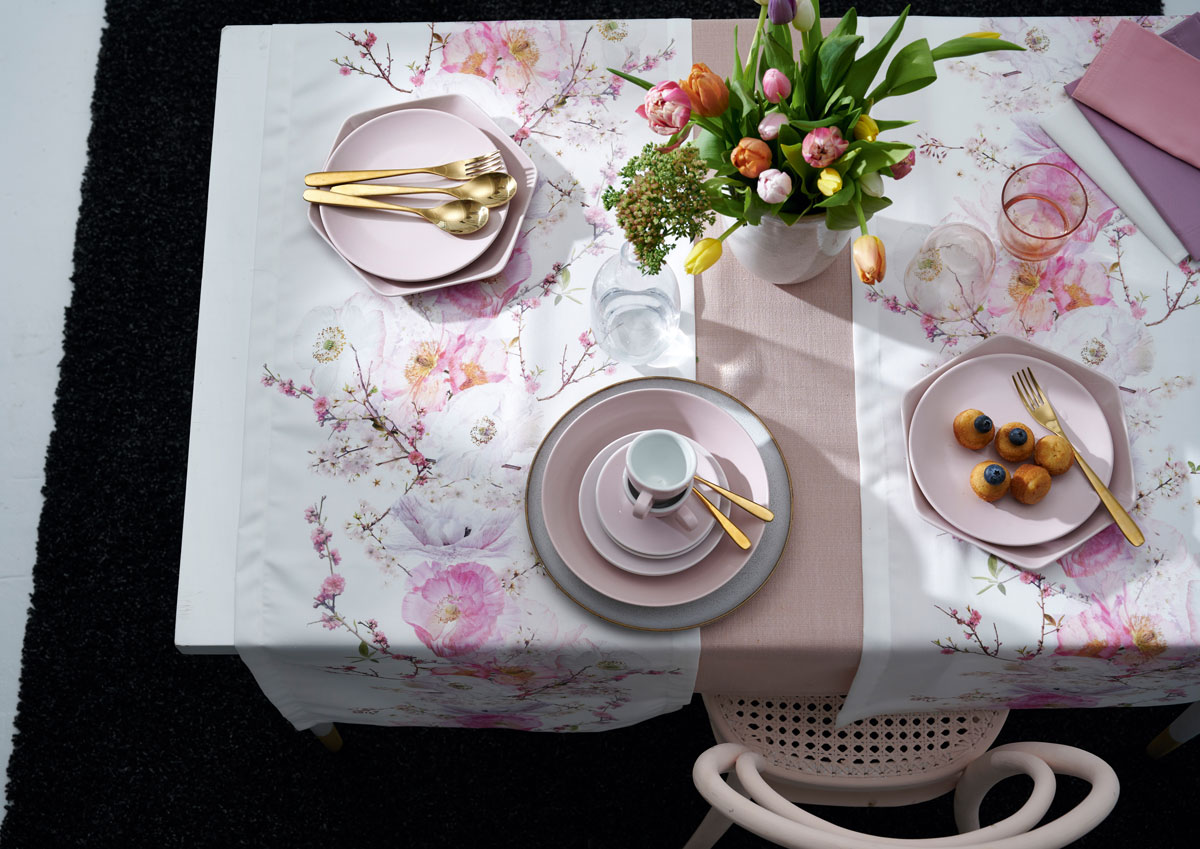 Gedeckter Tisch mit rosafarbenem und goldfarbenem Besteck, Tischläufer weiß mit rosa Blütendekor, weiße Vase mit bunten Tulpen