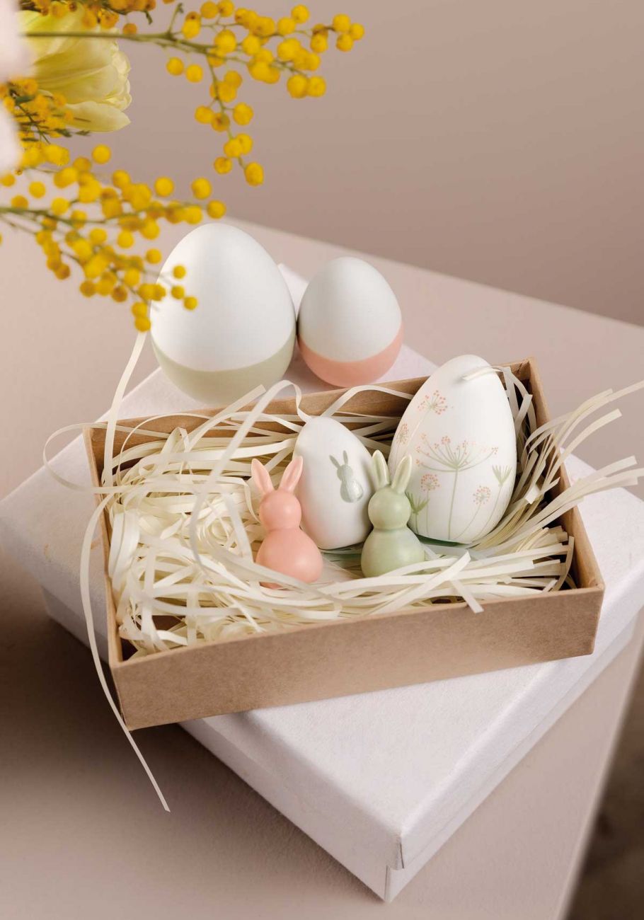 Geschenkekiste gefüllt mit Holzwolle, kleinen Osterhasen aus Porzellan und Wachskerzen in Eiform