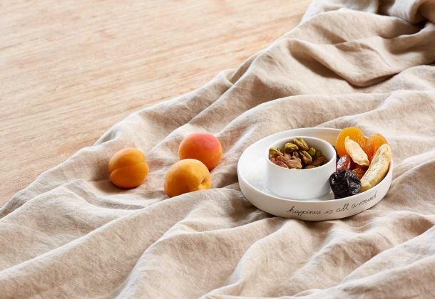 auf beigem Leinentuch befinden sich  Pfirsiche und eine weiße Prozellanschale mit getrocknetem Obst